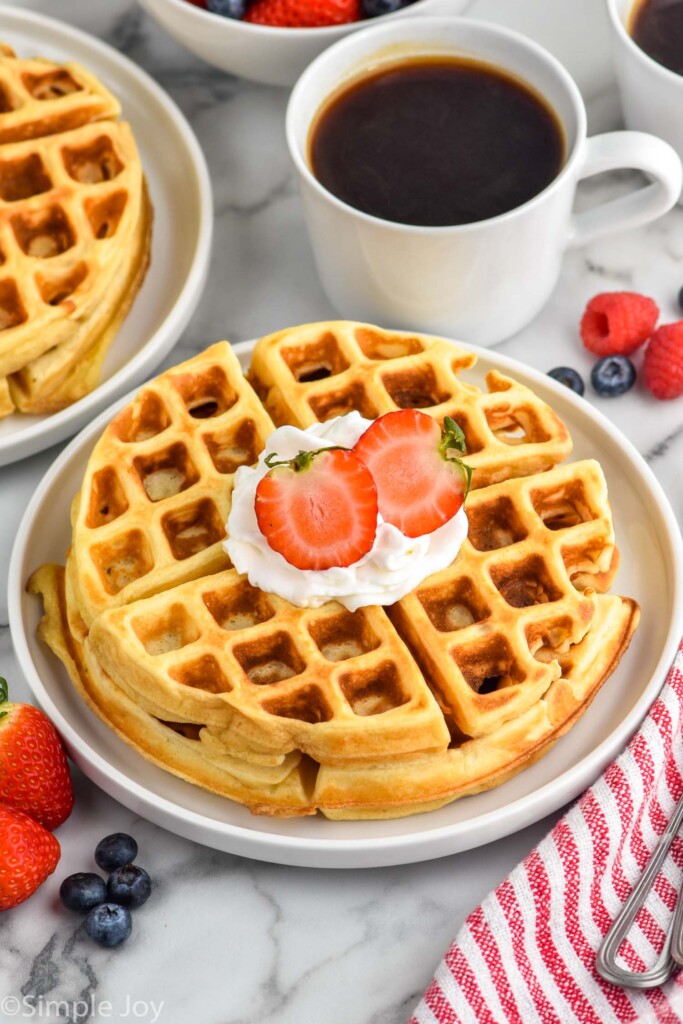 Photo of Buttermilk Waffles on a plate garnished with whipped cream and strawberries. Cup of coffee and more berries beside.