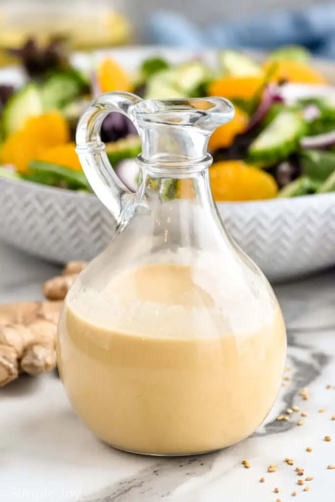 Jar of ginger dressing. Bowl of salad and fresh ginger sitting in background.