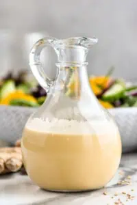 Jar of ginger dressing. Bowl of salad and fresh ginger sitting in background.