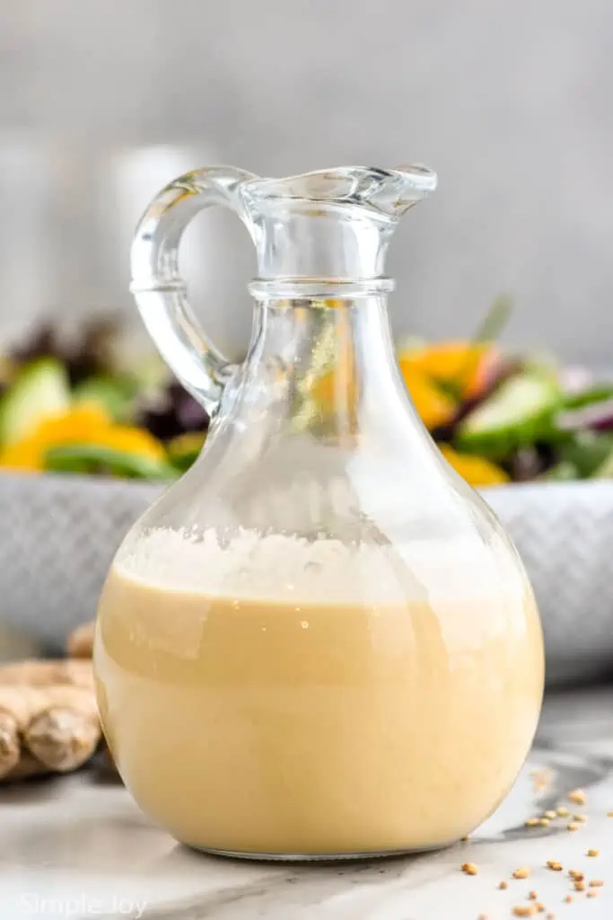 Jar of ginger dressing. Bowl of salad and fresh ginger sitting in background.