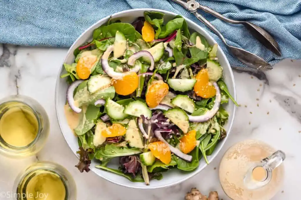 overhead of bowl of salad with ginger dressing. Ginger dressing ingredients and tongs sit beside.