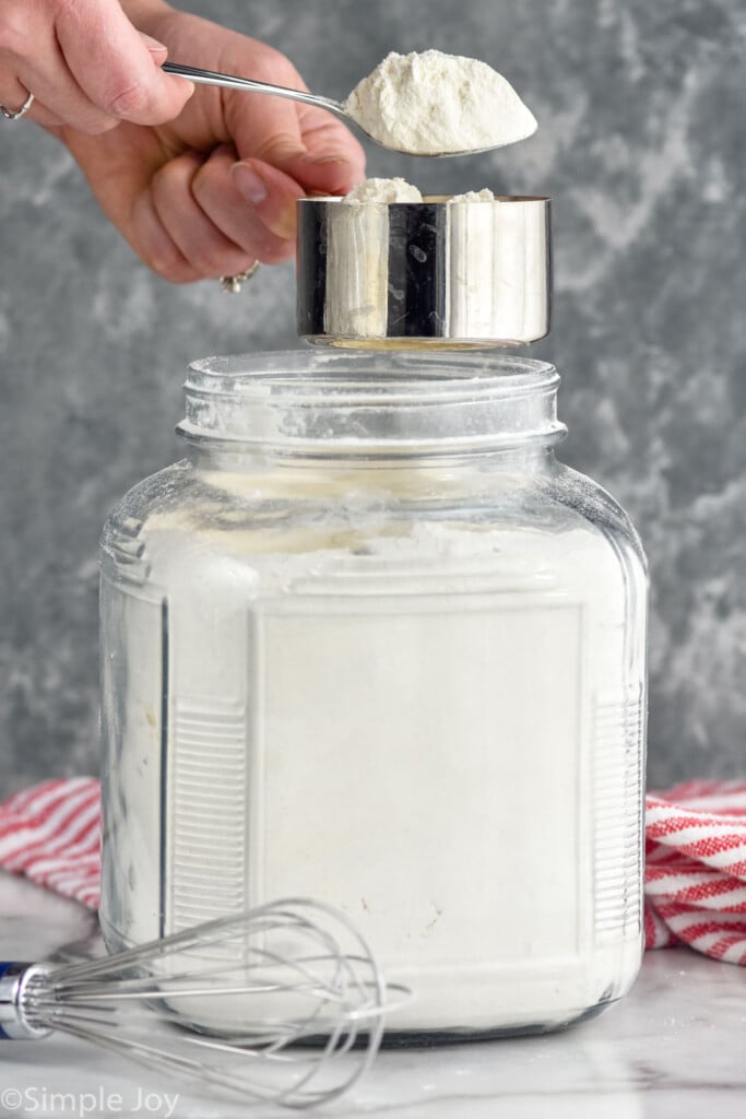 Person's hands spooning flour into measuring cup over flour container with whisk beside for How to Measure Flour