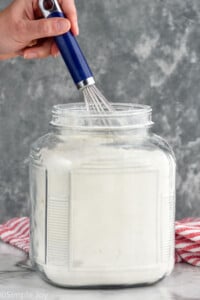 Person's hand holding whisk in flour container for How to Measure Flour