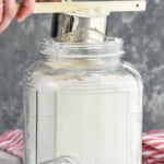 Person's hands leveling off measuring cup of flour with handle of spatula over container of flour with whisk beside for How to Measure Flour