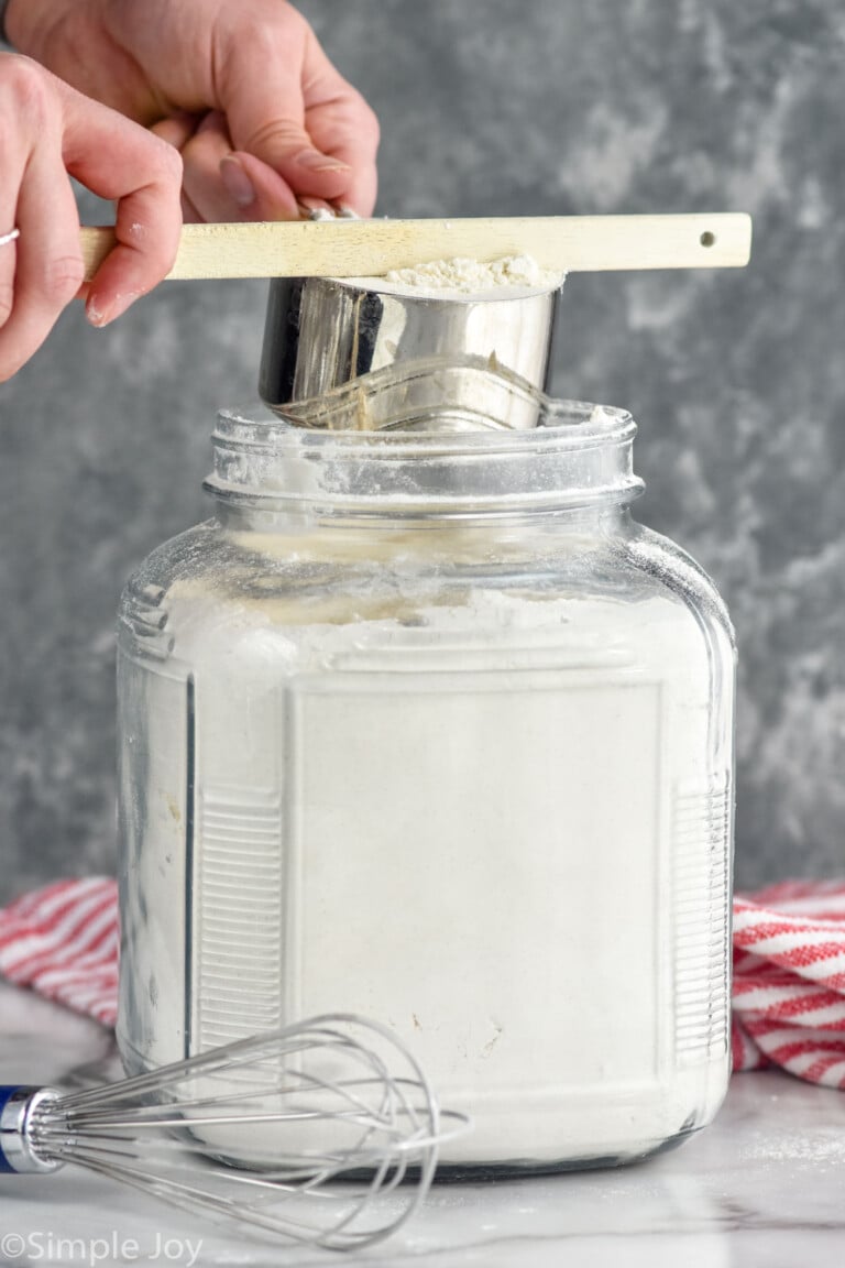 Person's hands leveling off measuring cup of flour with handle of spatula over container of flour with whisk beside for How to Measure Flour