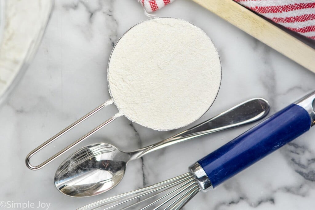 Overhead view of measuring cup of flour, spoon, and whisk for How to Measure Flour