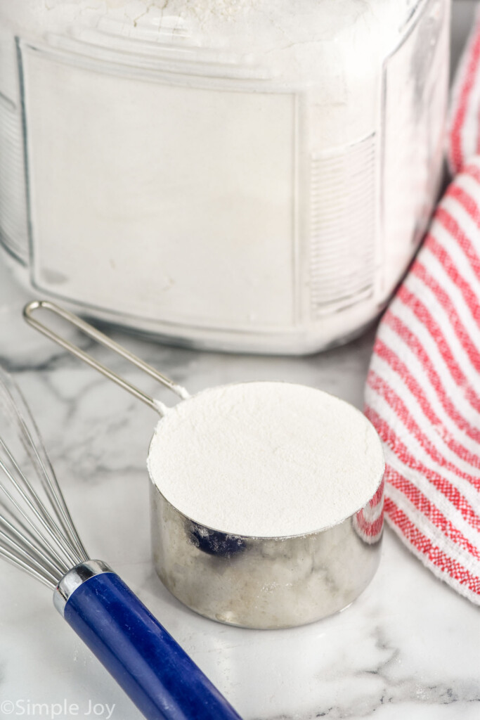 Measuring cup of flour with whisk and flour container beside for How to Measure Flour