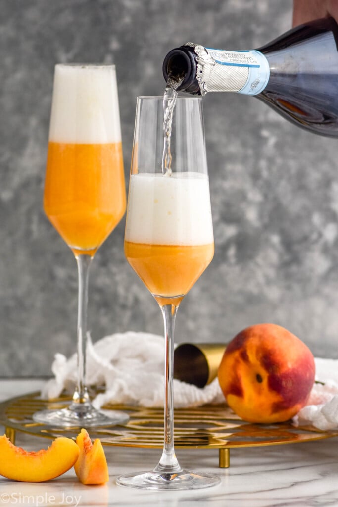 Side view of champagne being poured into champagne flute of ingredients for Bellinis recipe. peach and slices beside.