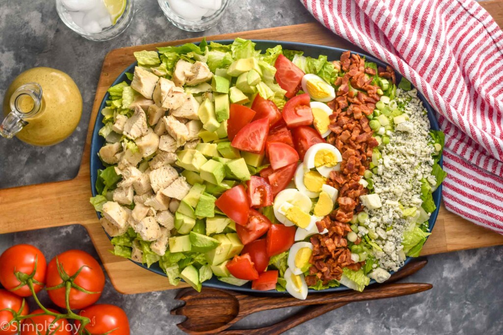Overhead photo of Cobb Salad with tomatoes, dressing, and salad tongs beside.
