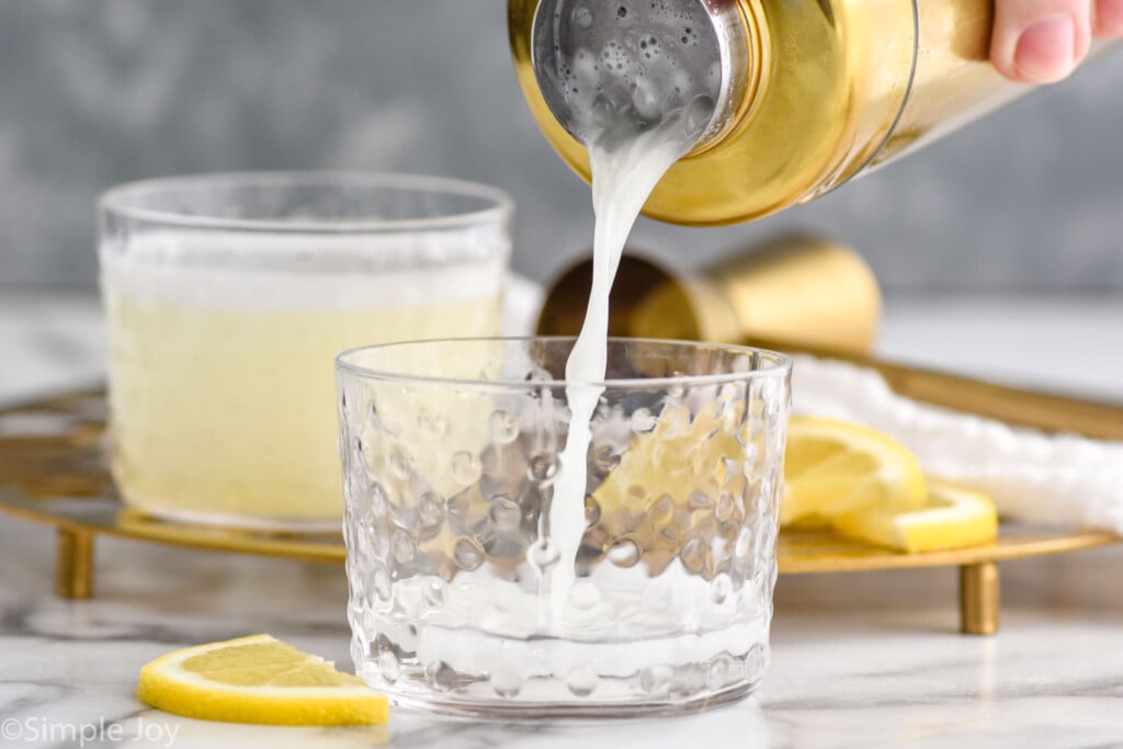 Side view of cocktail shaker of Gin Fizz recipe being poured into tumblers. Lemon slices and cocktail jigger beside.