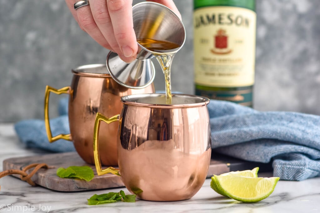 Side view of person's hand pouring Jameson into copper mugs for Irish Mule recipe, lime wedges and mint leaves beside. Jameson bottle beside.