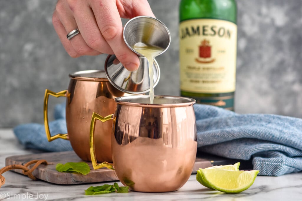 Person's hand pouring lime juice into copper mugs for Irish Mule recipe, bottle of Jameson, lime wedges, and mint leaves beside.