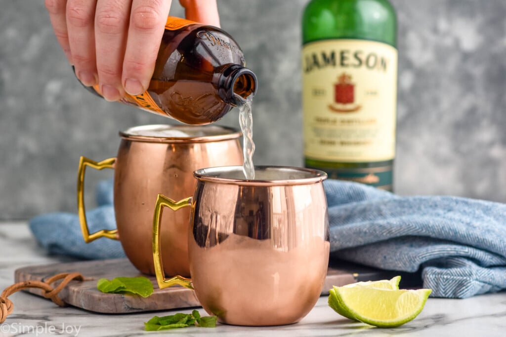 Side view of person's hand pouring ginger beer into copper mugs for Irish Mule recipe. Bottle of Jameson and lime wedges beside