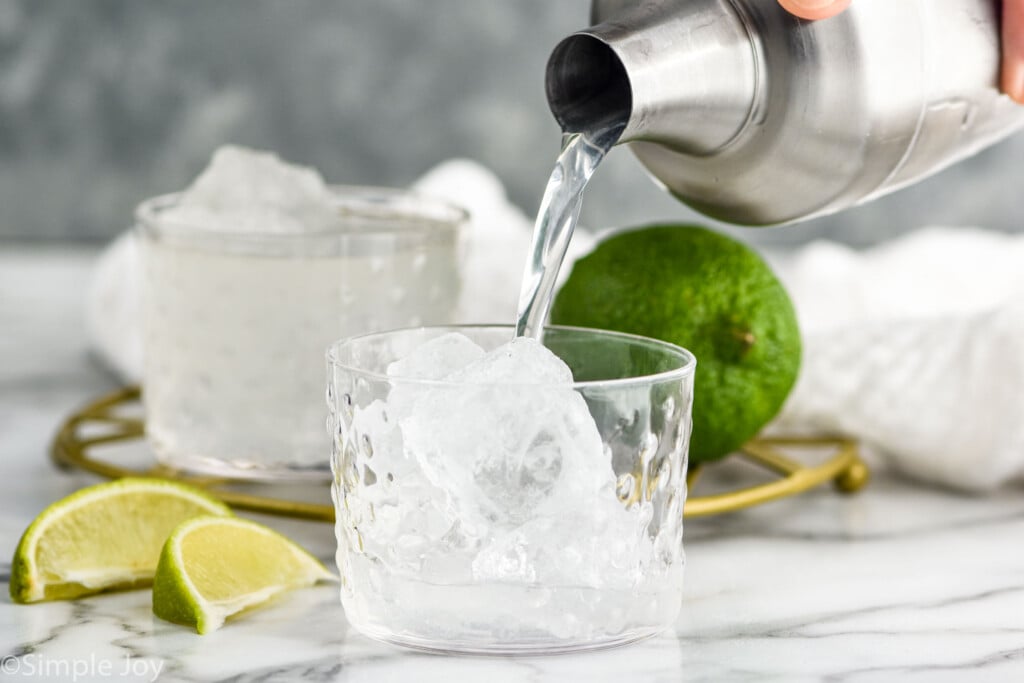 Side view of person's hand pouring shaker bottle of ingredients into tumbler of ice for Vodka Gimlet recipe. Another tumbler of Vodka Gimlet, lime wedges, and lime beside.