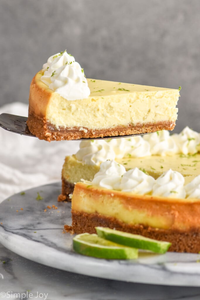 serving spatula lifting a slice of key lime pie cheesecake out of the cheesecake. Lime slices sitting beside.