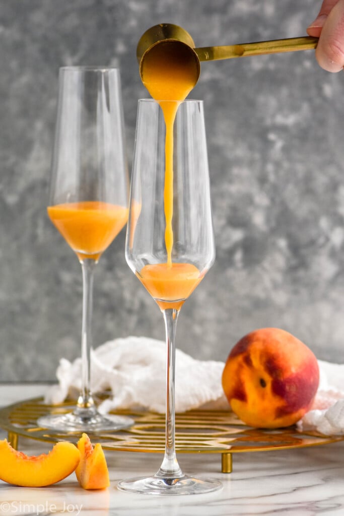 Side view of cocktail jigger pouring ingredients into champagne flutes for Bellinis recipe. Peach slices and peach beside.