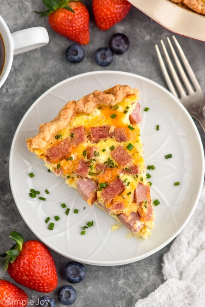overhead photo of a slice of Ham and Cheese Quiche on a plate with fork and fruit beside.