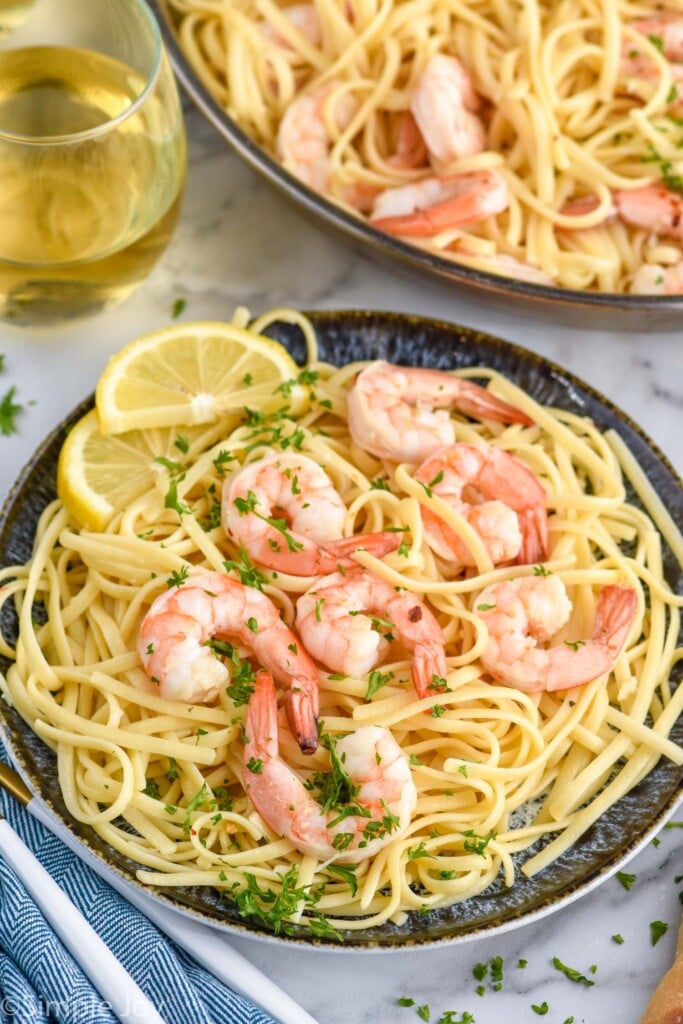 Plate of Shrimp Linguine with lemon wedges with skillet of Shrimp Linguine and glass of wine beside