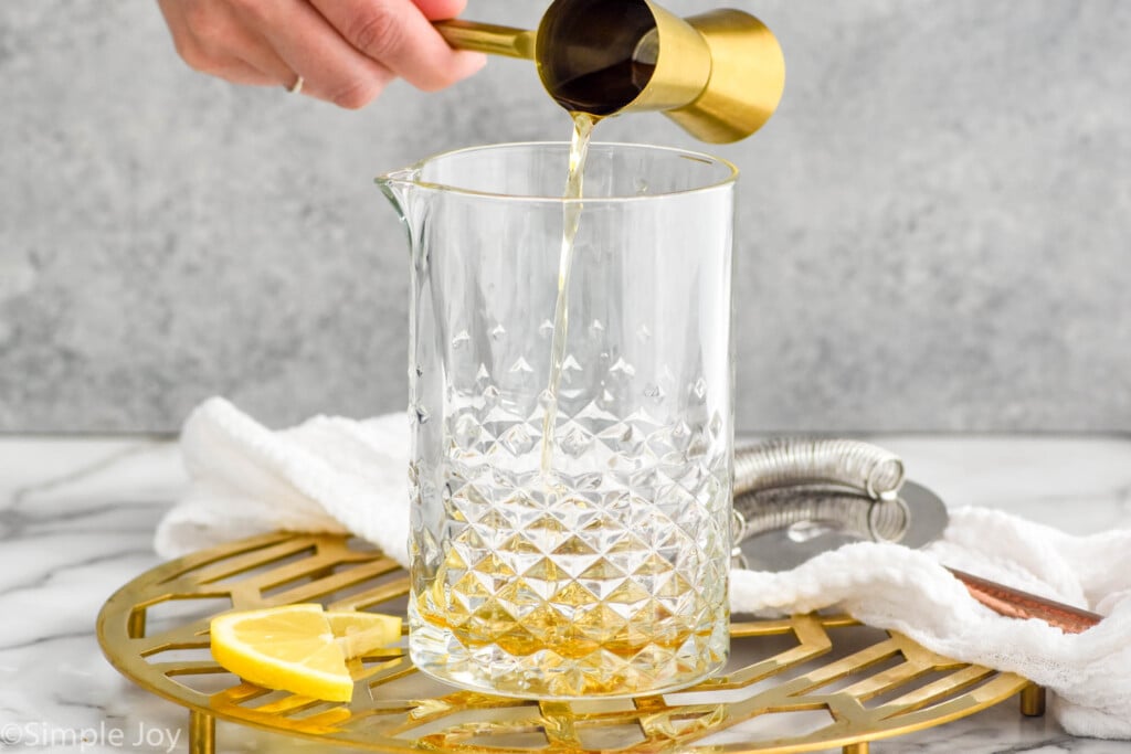 Side view of person's hand pouring cocktail jigger of ingredient into pitcher for Gold Rush Cocktail recipe. Lemon slices beside.