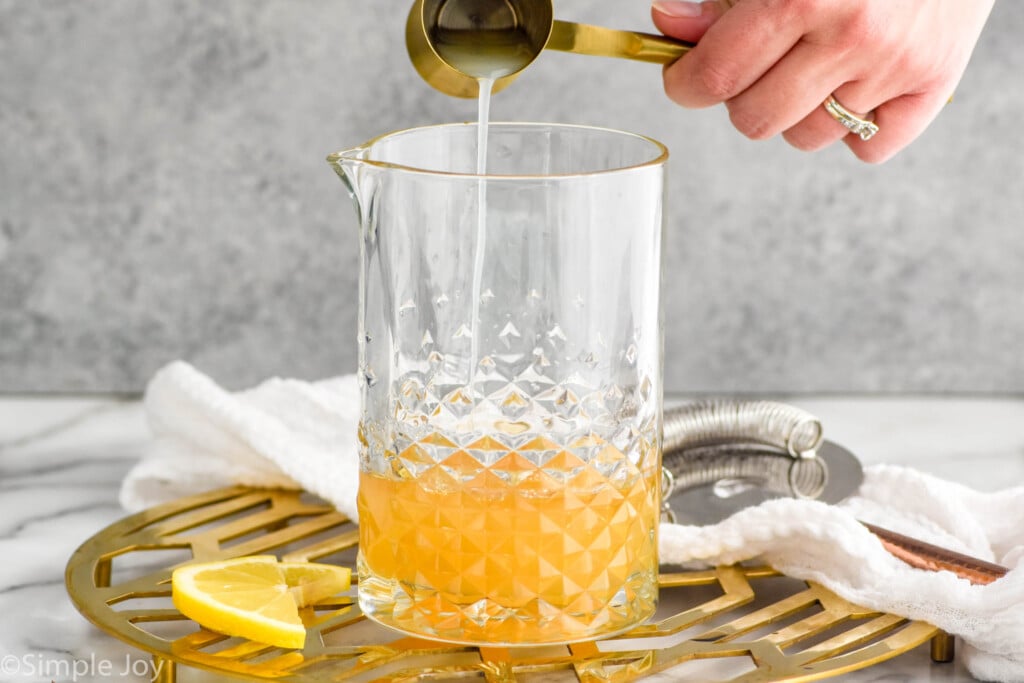 Side view of person's hand pouring cocktail jigger of lemon juice into pitcher of ingredients for Gold Rush Cocktail recipe. Lemon slices beside.