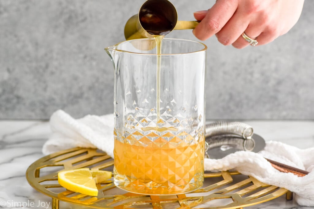 Side view of person's hand pouring cocktail jigger of honey into pitcher of ingredients for Gold Rush Cocktail recipe. Lemon slices beside.