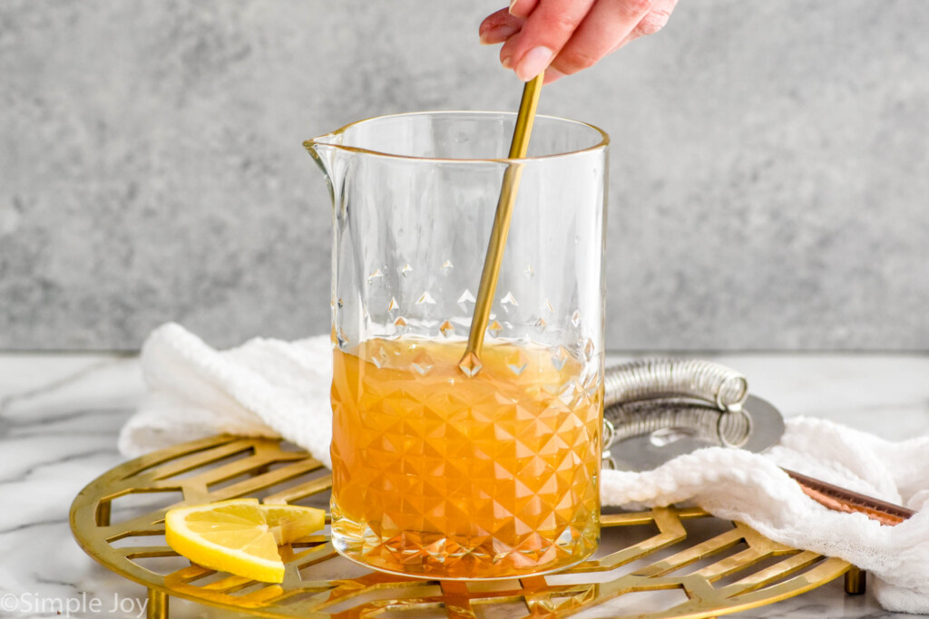 Side view of pitcher of Gold Rush Cocktail being stirred. Lemon slices beside.