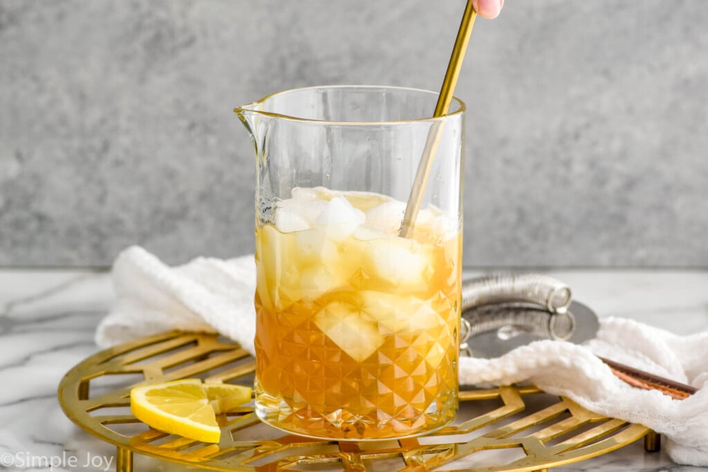 Side view of person's hand stirring pitcher of Gold Rush Cocktail recipe with ice. Lemon slices beside.