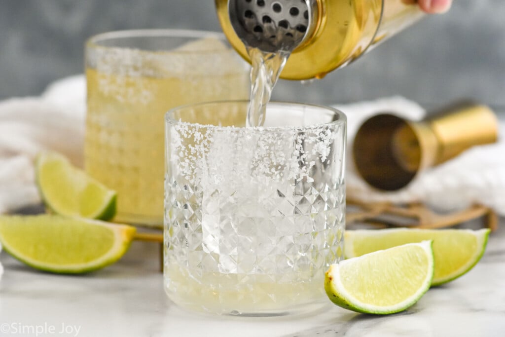 Side view of cocktail shaker of Margarita being poured into tumbler with salted rim, lime wedges beside.