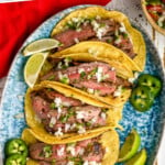 pinterest graphic of overhead of a plate of carne asada meat in tacos, topped with cojito cheese, onions, and jalapeños, says "the best carne asada simplejoy.com"