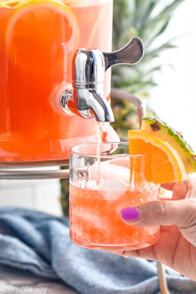 woman's hand holding glass of ice and rum punch garnished with slice of orange and slice of pineapple while dispenser of rum punch pours into glass.