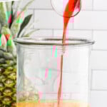 pinterest graphic for rum punch. Text says "the best rum punch recipe simplejoy.com" Image shows grenadine pouring into drink dispenser of rum punch ingredients. Fresh pineapple and orange slices sitting in background.