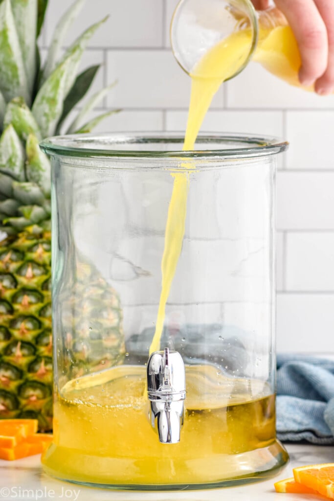 orange juice pouring into a drink dispenser of rum punch ingredients to make rum punch. Fresh pineapple and orange slices sitting in background