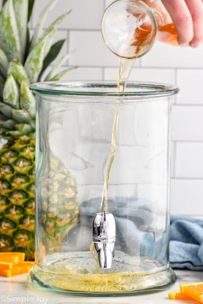 rum pouring into a drink dispenser to make rum punch. Fresh pineapple and orange slices sitting in background