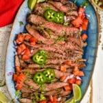 overhead of a serving dish with grilled and cut up carne asada garnished with fresh jalapeños, tomatoes, and lime wedges