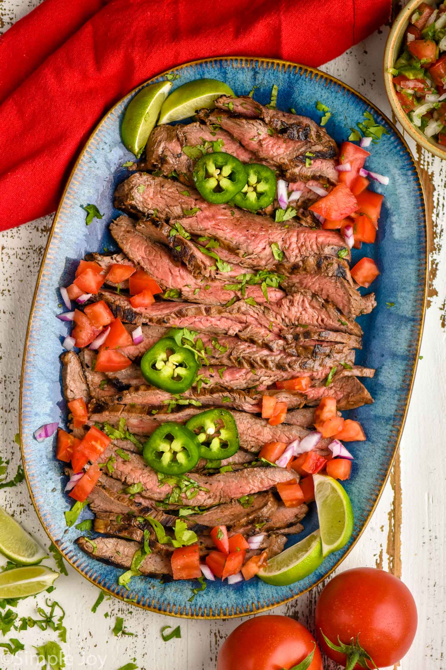 overhead of a serving dish with grilled and cut up carne asada garnished with fresh jalapeños, tomatoes, and lime wedges