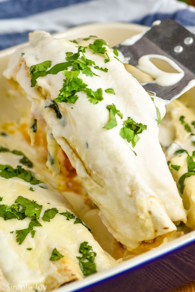 two chicken enchiladas with sour cream sauce being dished out of a casserole dish, garnished with cilantro