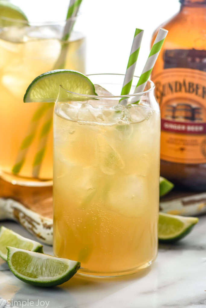 glass of Dark and Stormy with ice, straws, and a lime. Bottle of ginger beer, lime wedges, and glass of dark and stormy sitting in background.