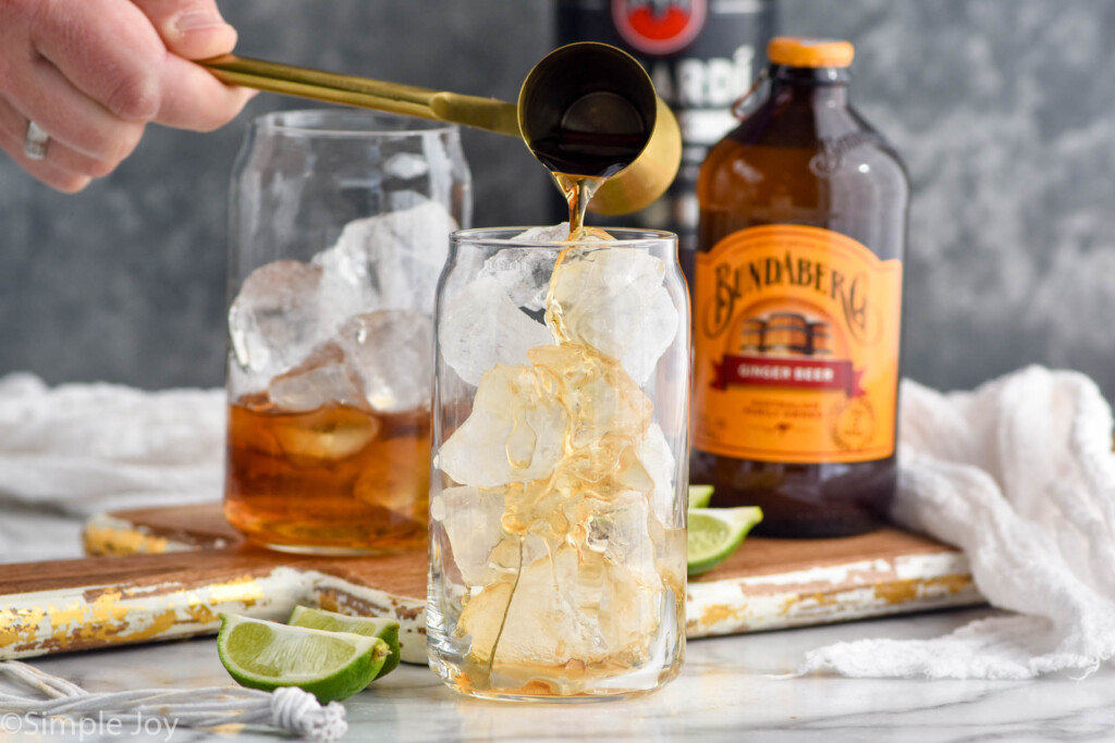 man's hand pouring cocktail jigger of dark rum into a glass of ice to make a dark and stormy. Dark and stormy ingredients sitting in background.