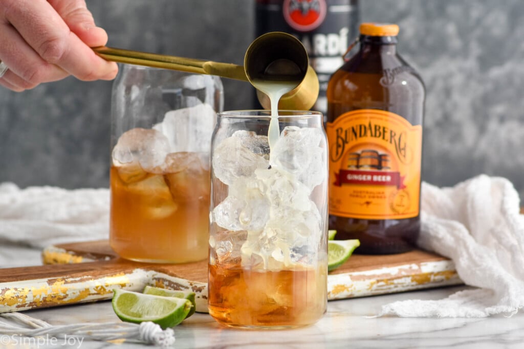 Man's hand pouring cocktail jigger of lime juice into a glass of dark and stormy ingredients. Lime wedges, ginger beer, and rum sitting in background.