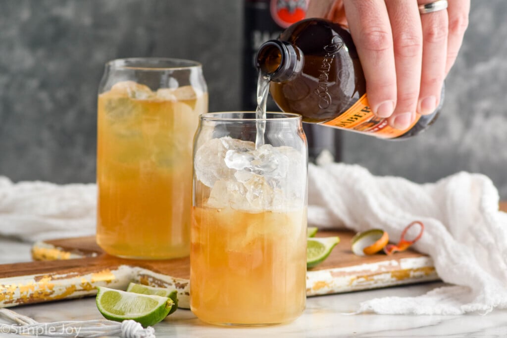 man's hand pouring bottle of ginger beer into a glass of dark and stormy ingredients and ice