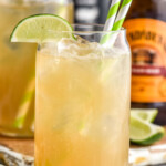 Glass of dark and stormy with ice, two straws, and a lime wedge garnish. Glass of dark and stormy, lime wedges, and bottle of ginger beer sitting in background. - 9