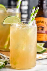 Glass of dark and stormy with ice, two straws, and a lime wedge garnish. Glass of dark and stormy, lime wedges, and bottle of ginger beer sitting in background.