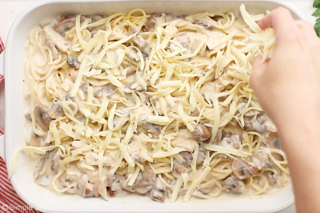 overhead of baking dish of Chicken Tetrazzini with person's hand sprinkling shredded cheese on top