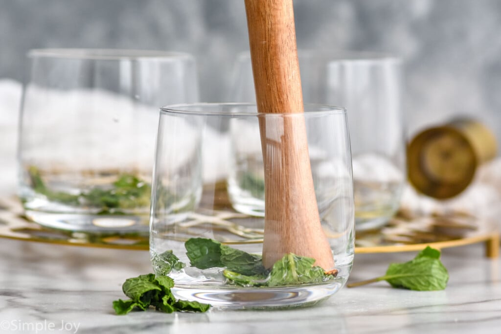 cocktail muddler muddling mint in a glass showing how to make a mint julep. Two glasses and fresh mint leaves sitting in background