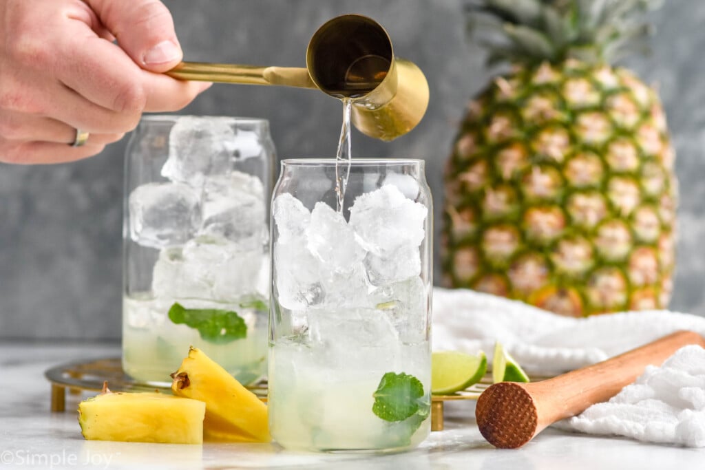 Side view of cocktail jigger being poured into glass of ice and ingredients for Pineapple Mojito recipe.