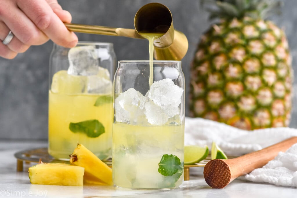 Side view of cocktail jigger being poured into glass of ice and ingredients for Pineapple Mojito recipe. Pineapple chunks and lime wedges beside, along with whole pineapple and cocktail muddler.