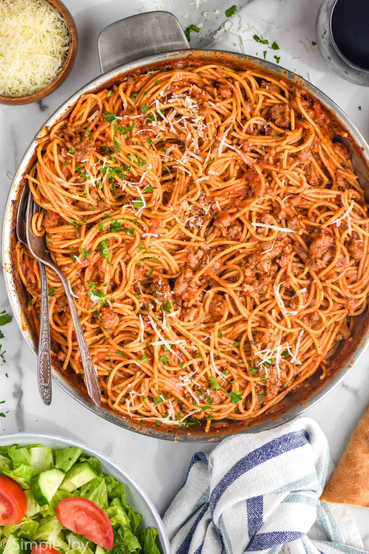 Overhead view of a pot of Sausage Spaghetti in large skillet garnished with shredded parmesan and parsley, two forks for serving. with salad, bread, red wine, and parmesan beside.