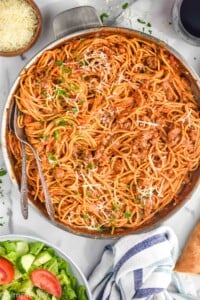 Overhead view of a pot of Sausage Spaghetti in large skillet garnished with parsley and shredded parmesan cheese with salad and parmesan beside. Two forks in skillet for serving.