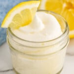 jar of Lemon Buttercream Frosting with a slice of lemon on top. Slices of lemon sitting in front and behind