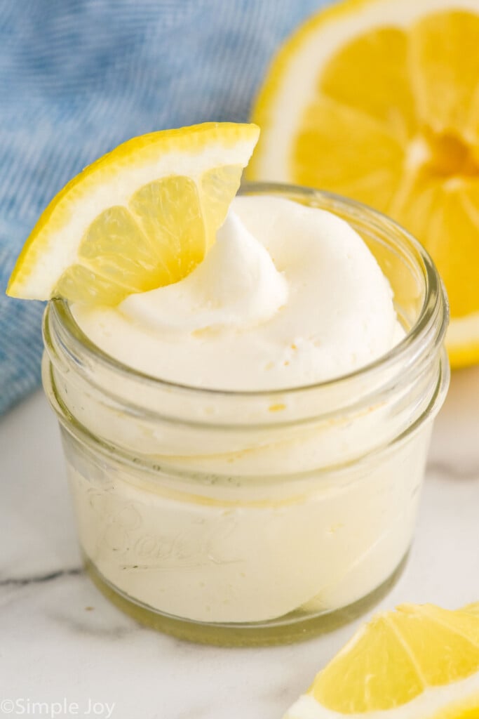 jar of Lemon Buttercream Frosting with a slice of lemon on top. Slices of lemon sitting in front and behind